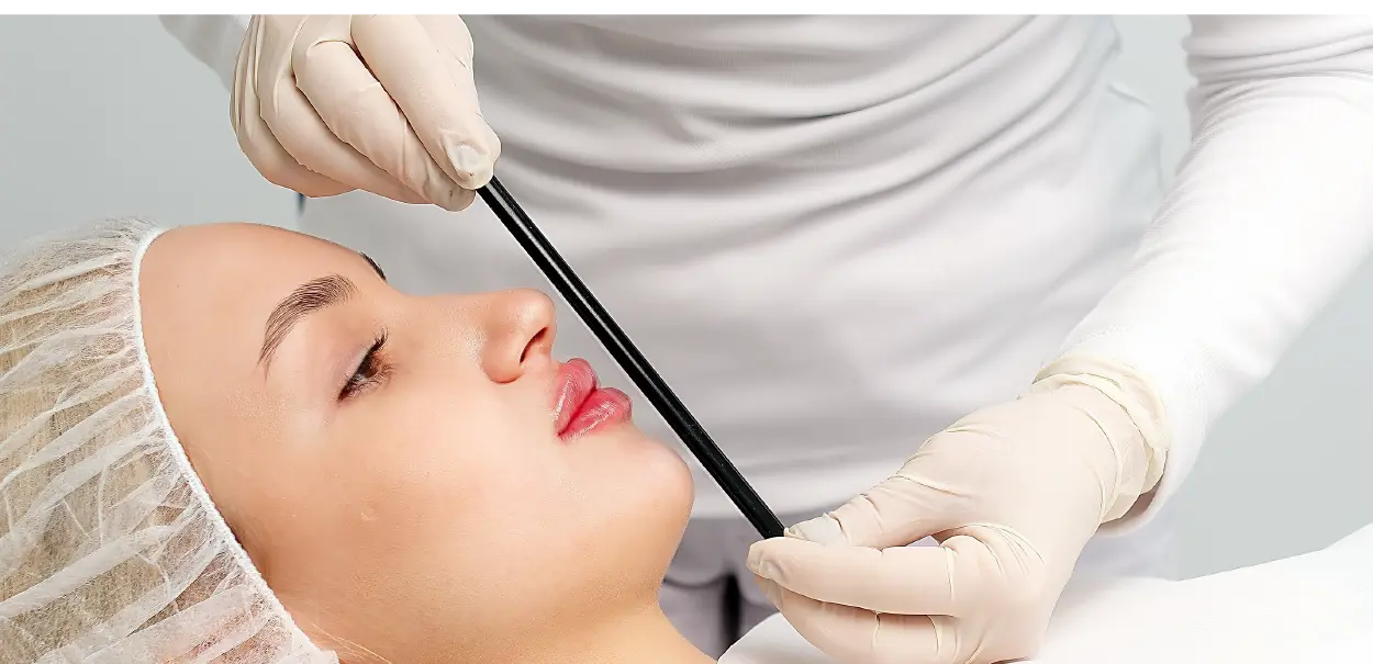 A person with long hair wearing a hairnet lying down while a figure in gloves holds a black rod next to their mouth, suggesting a dental or cosmetic procedure.
