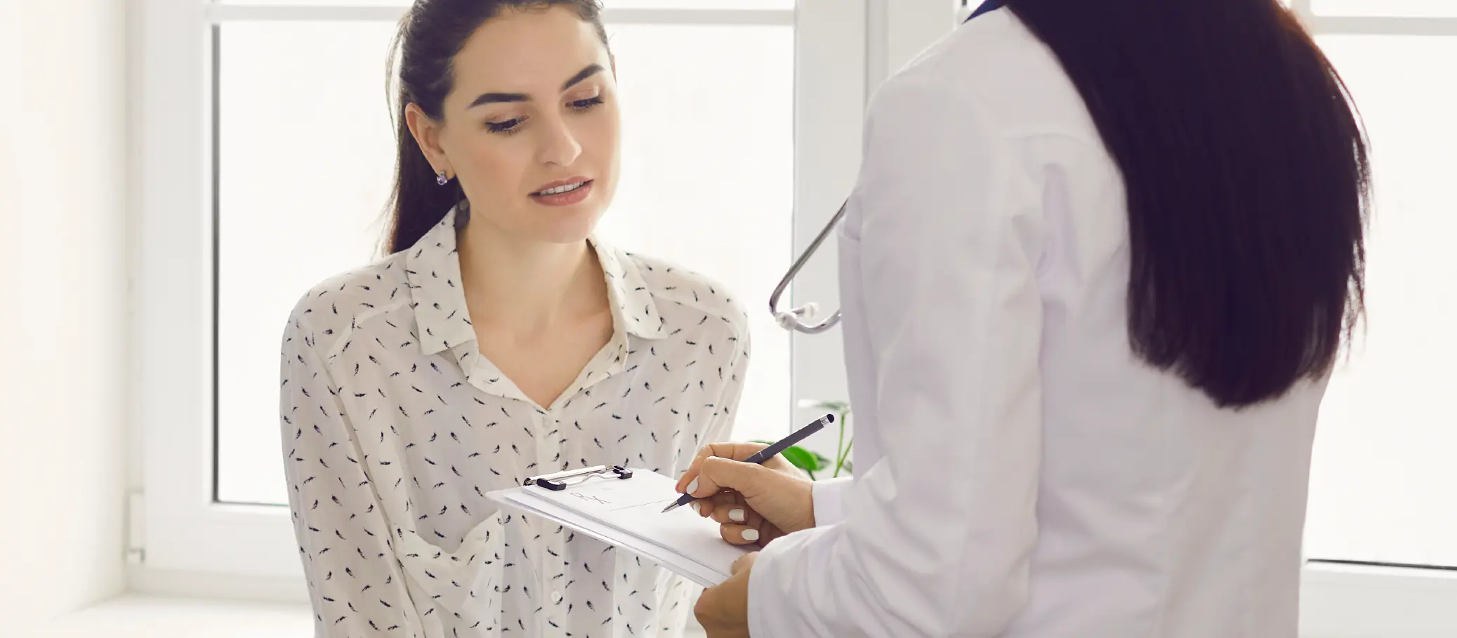 A female doctor in a white coat holding a clipboard and pen while examining a female patient who is wearing a white blouse with a pattern of small black lines.