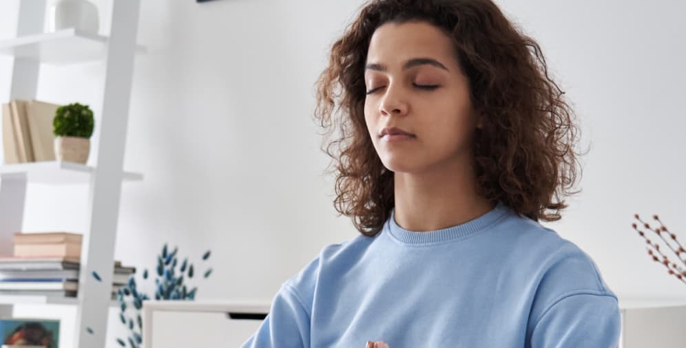 A young woman with curly hair, wearing a light blue sweatshirt, sitting with her eyes closed in a meditative pose.