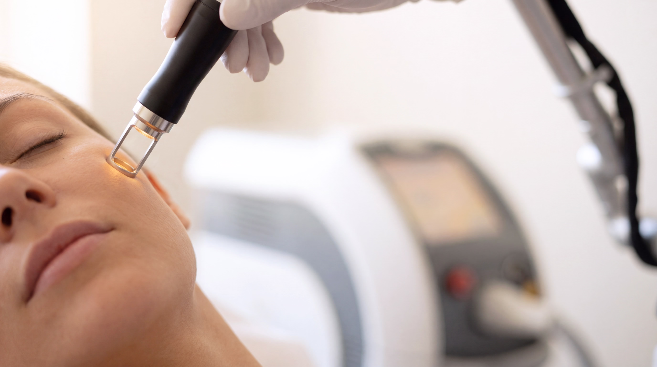 A hand holding a black device with a transparent tip, positioned near the cheek of a woman receiving a facial treatment.