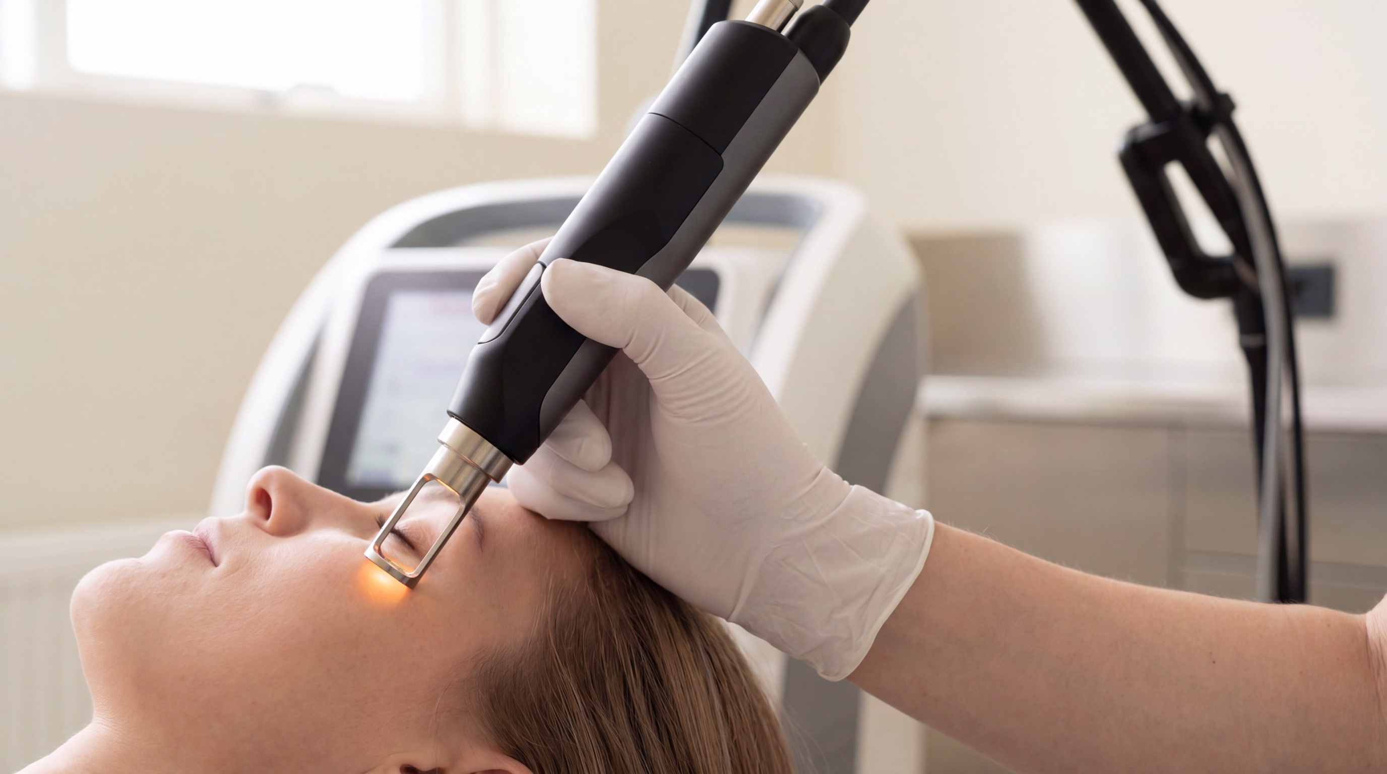 A hand wearing a white glove holding a handheld laser device pointing towards the forehead of a person lying down. The device emits a small orange light at the tip.