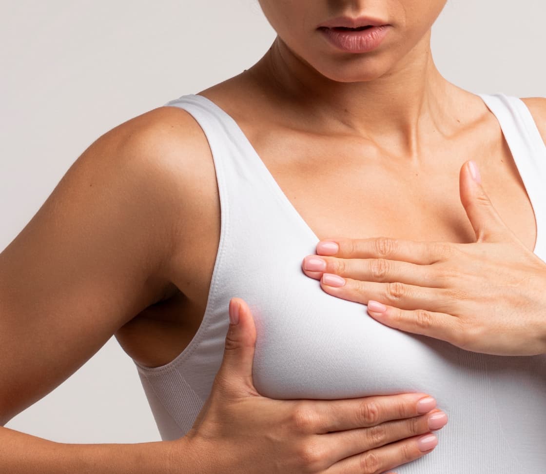 A person wearing a white tank top using both hands to cover their chest, with a serious expression on their face.
