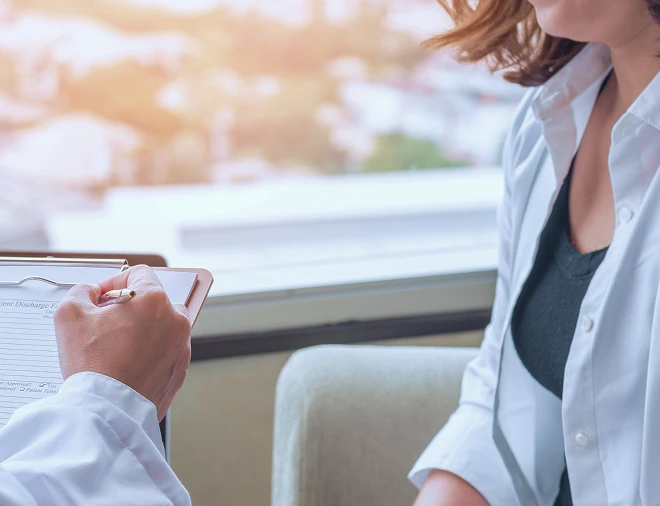 A medical professional in a white coat taking notes on a clipboard while seated next to a patient wearing a black top.