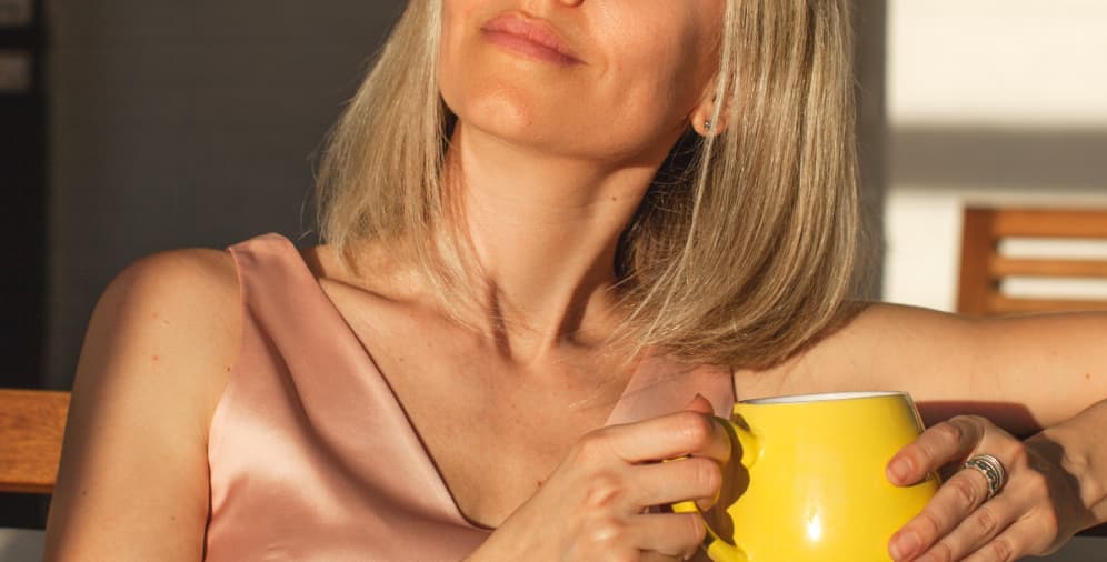 A woman with medium-length blonde hair wearing a pink satin top, holding a yellow mug, and looking thoughtfully towards the light.