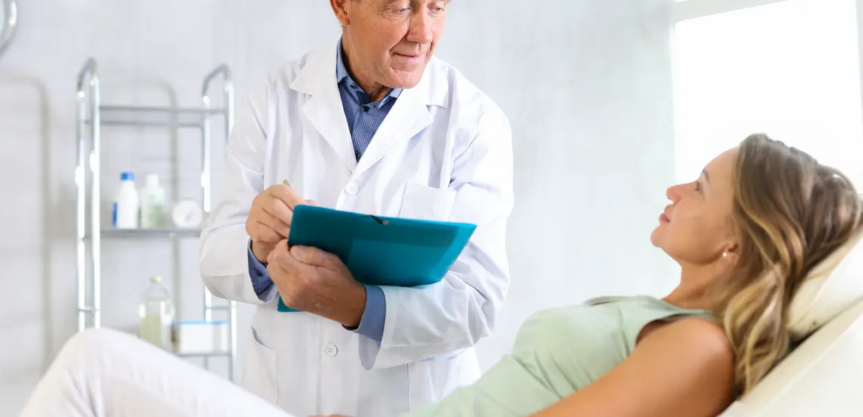 A doctor in a white lab coat holding a clipboard is speaking with a patient lying on an examination table.