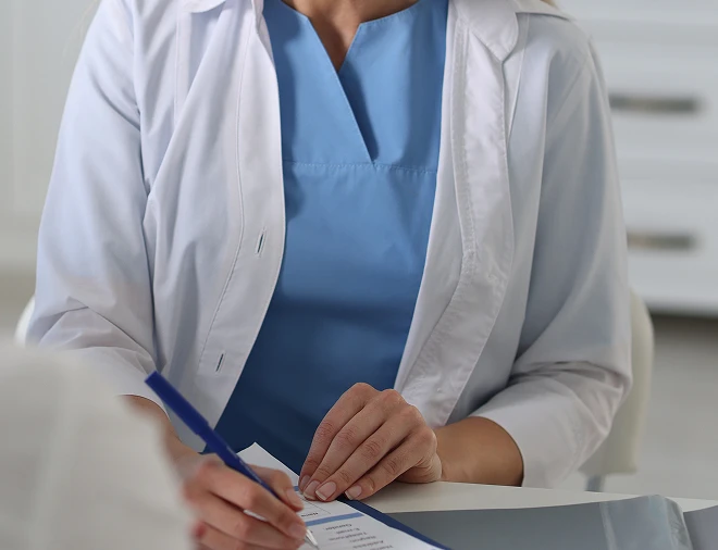 A person in a light blue medical scrub and a white lab coat is holding a pen and writing on a clipboard.