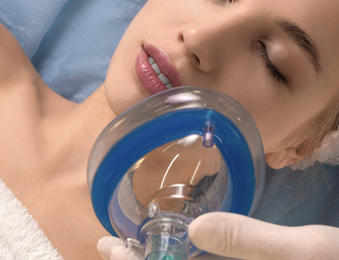 A woman with closed eyes lying on a treatment table, holding a clear medical mask with a blue rim, while a gloved hand supports the mask. Her skin appears smooth and healthy.