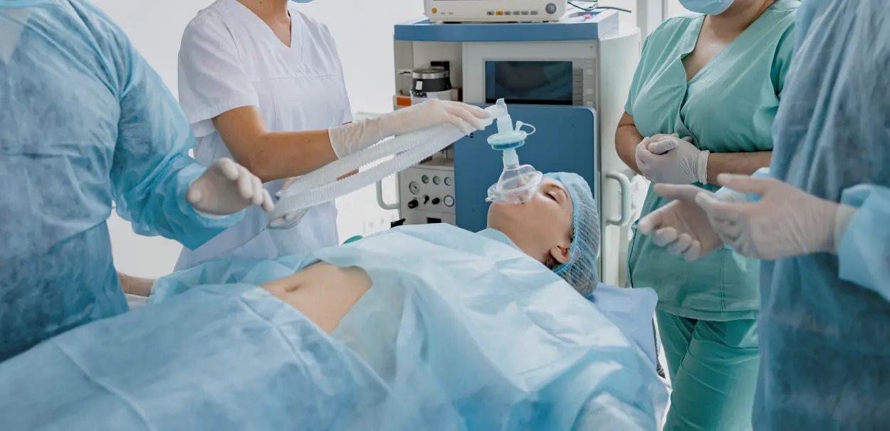 A patient lying on an operating table while medical staff in scrubs administer anesthesia using a mask.