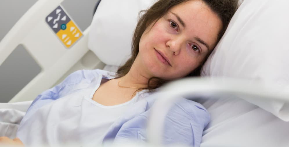 A woman lying in a hospital bed, looking towards the camera with a neutral expression. She has brown hair and is wearing a light blue hospital gown. The bed is equipped with white bedding and a hospital rail.