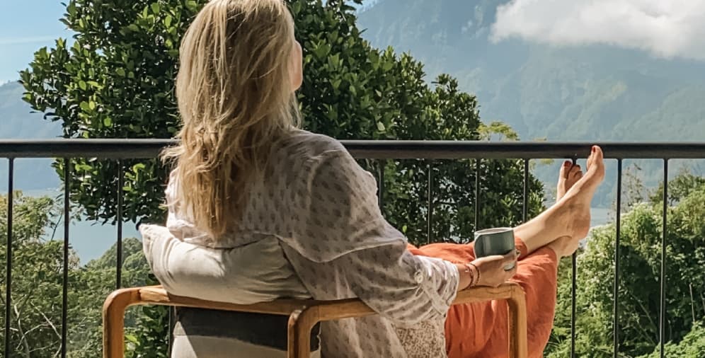 A woman sitting on a wooden chair with her feet up, holding a small cup. She has long, blonde hair and is wearing a loose white shirt and orange pants. The background features lush greenery and mountains.