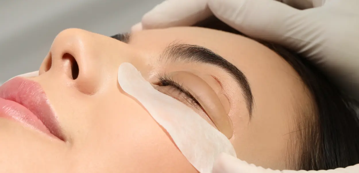 A close-up of a woman's face receiving a beauty treatment, with a clear gel patch placed under her left eye. A gloved hand gently holds her forehead.