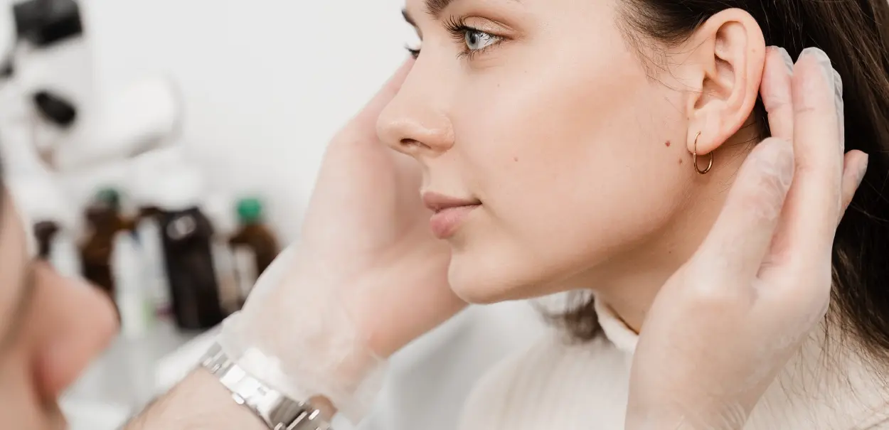 A profile view of a woman with light skin and earrings, receiving a cosmetic treatment from a person wearing a glove. The woman is looking slightly upwards, and her hair is neatly styled.