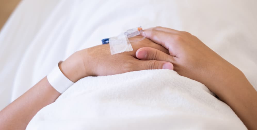 A person's hand resting on another hand, both partially covered by a white sheet, with an IV catheter attached to the wrist of the hand on top.