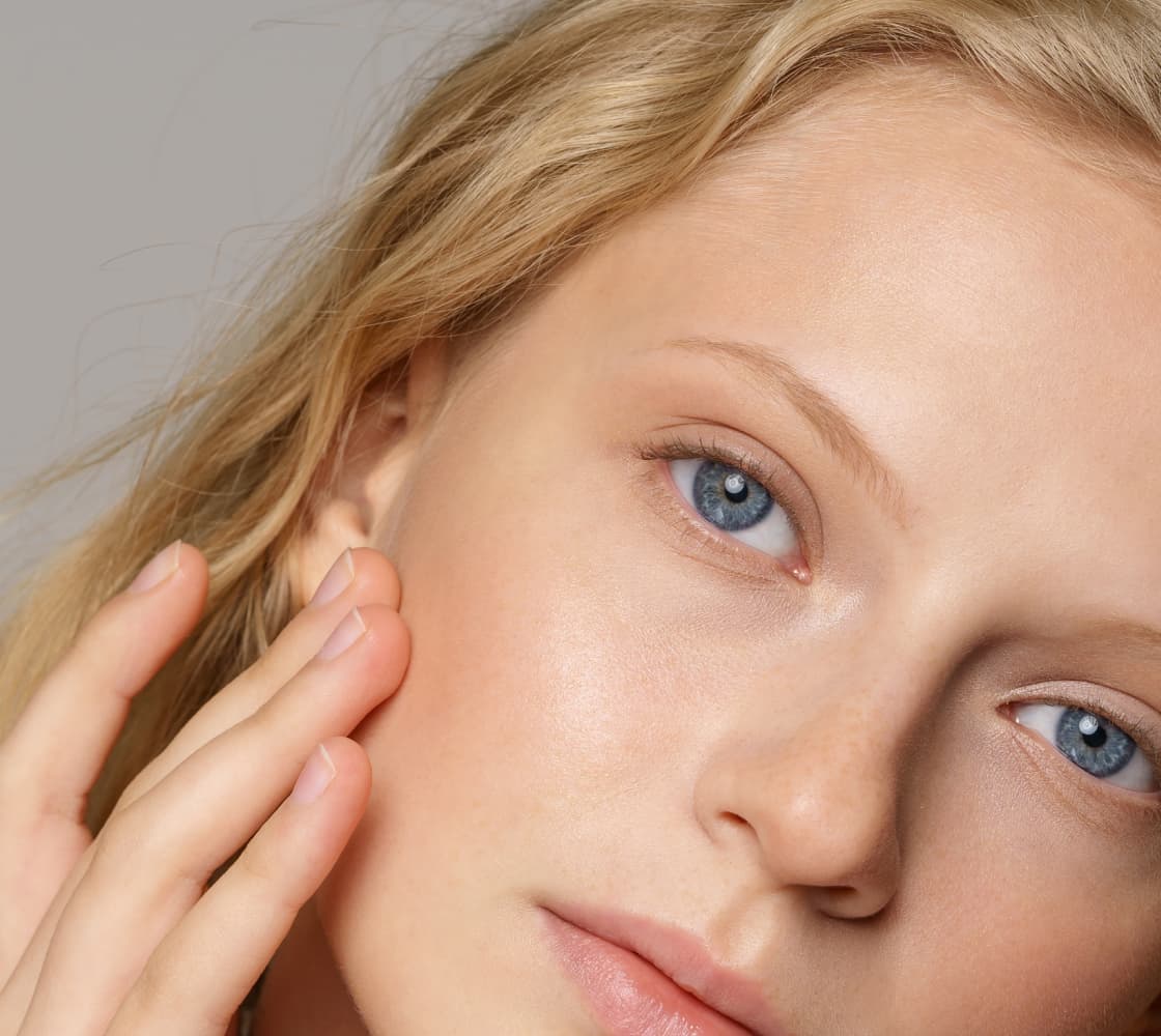 A close-up of a young woman with blonde hair, blue eyes, and natural makeup, gently touching her cheek.