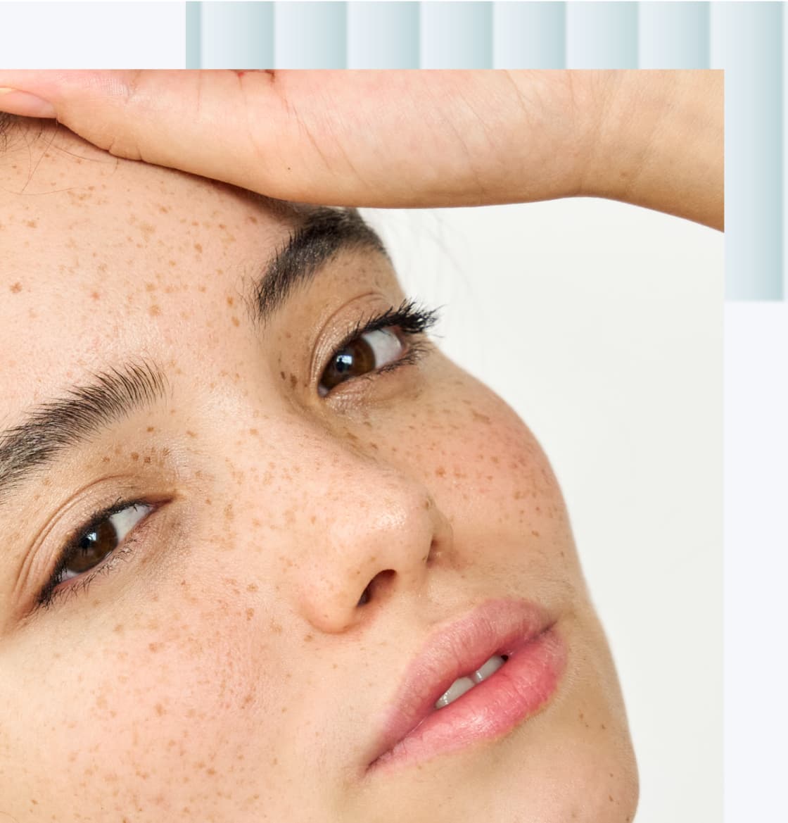 A close-up of a young woman with freckles, gently touching her forehead with one hand, displaying a natural makeup look and soft, subtle lipstick.