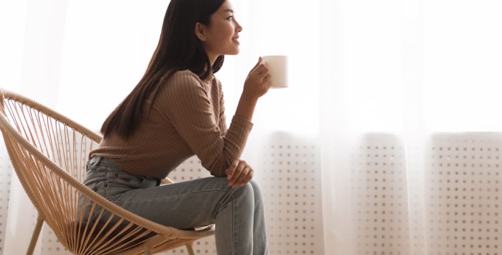 A woman sitting in a rattan chair, holding a white mug and looking contemplatively to the side.