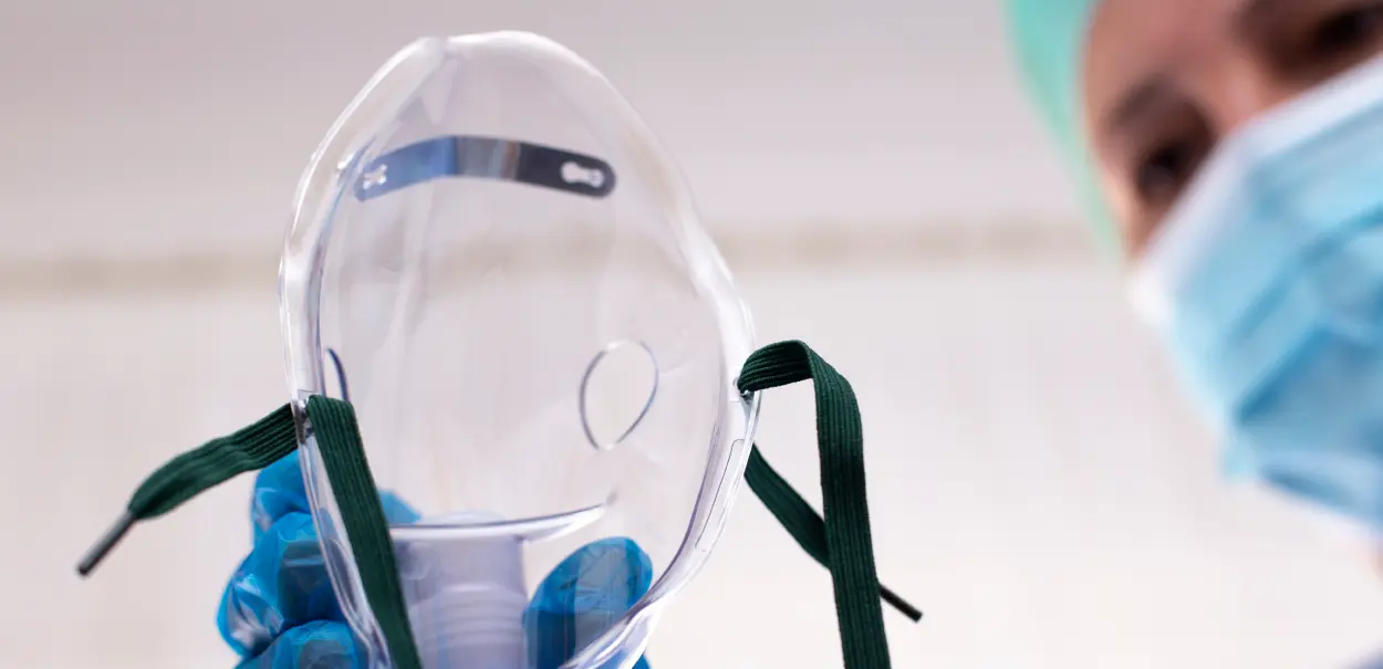 A person wearing blue gloves holds a clear medical face mask with two green straps.