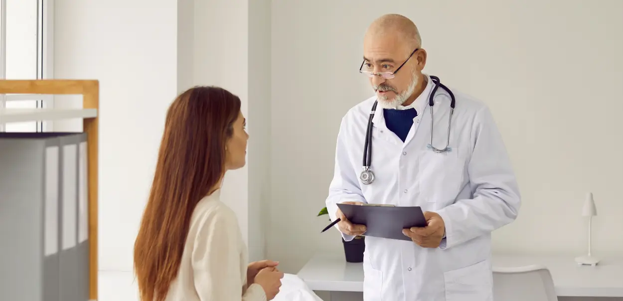 A doctor in a white coat with a stethoscope around his neck, holding a clipboard and speaking to a patient.