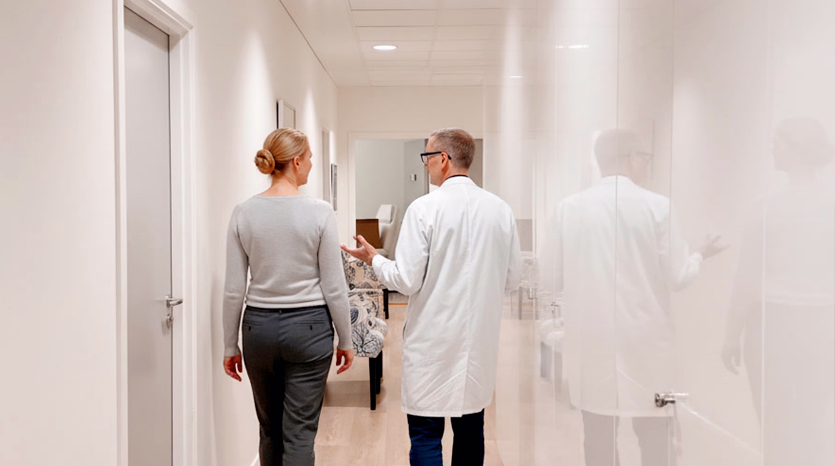 A doctor in a white coat talking to a patient walking down a well-lit corridor with a modern and clean aesthetic.