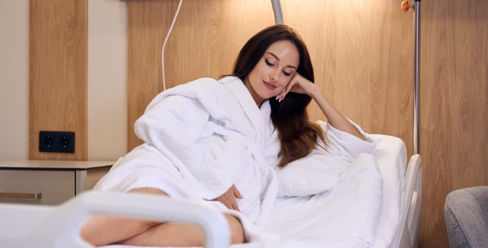 A woman in a white robe sitting on a hospital bed, resting her chin on her hand and smiling softly.
