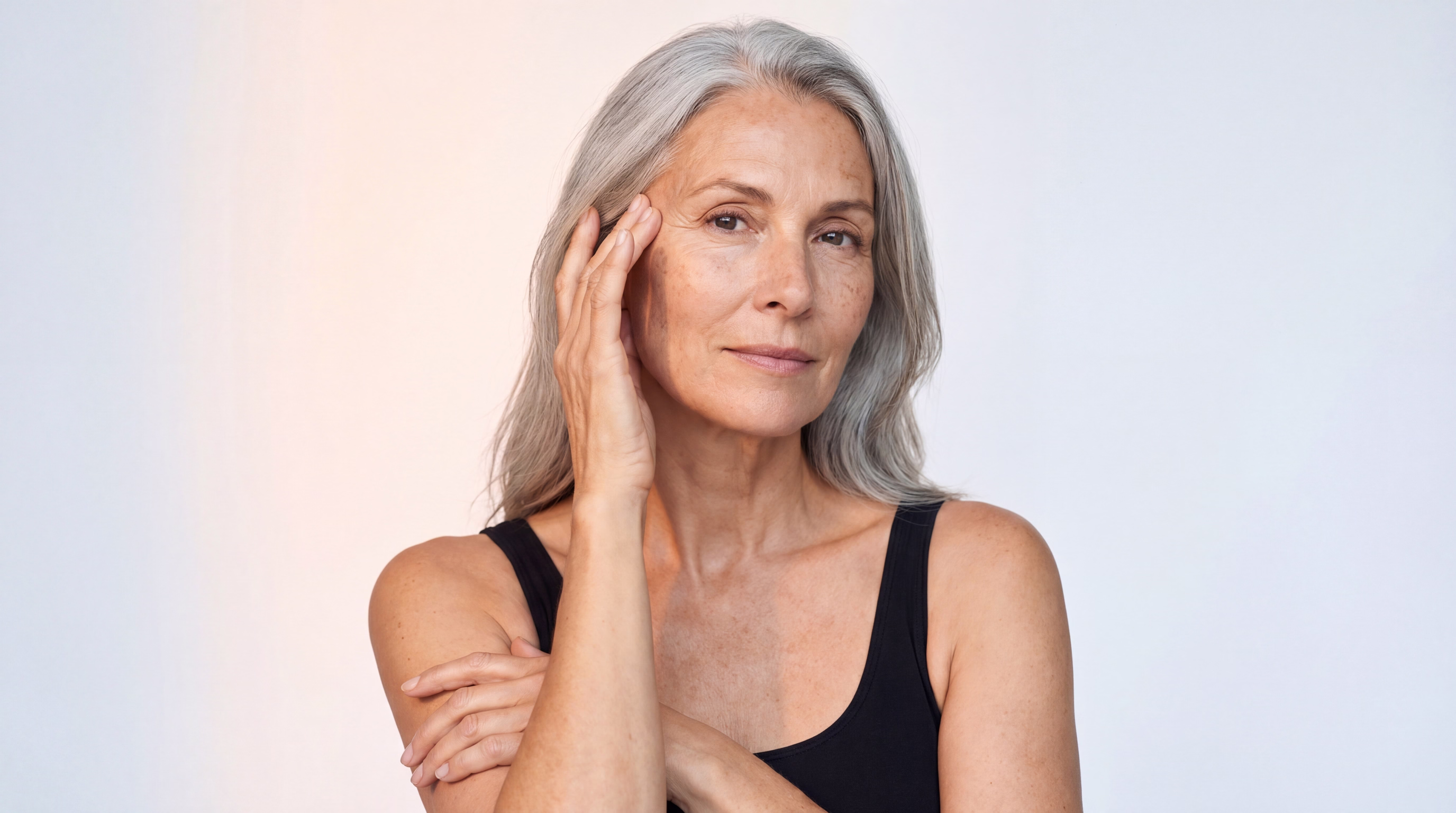 A mature woman with long, gray hair, gently touching her face with one hand and resting the other against her arm, wearing a black sleeveless top.