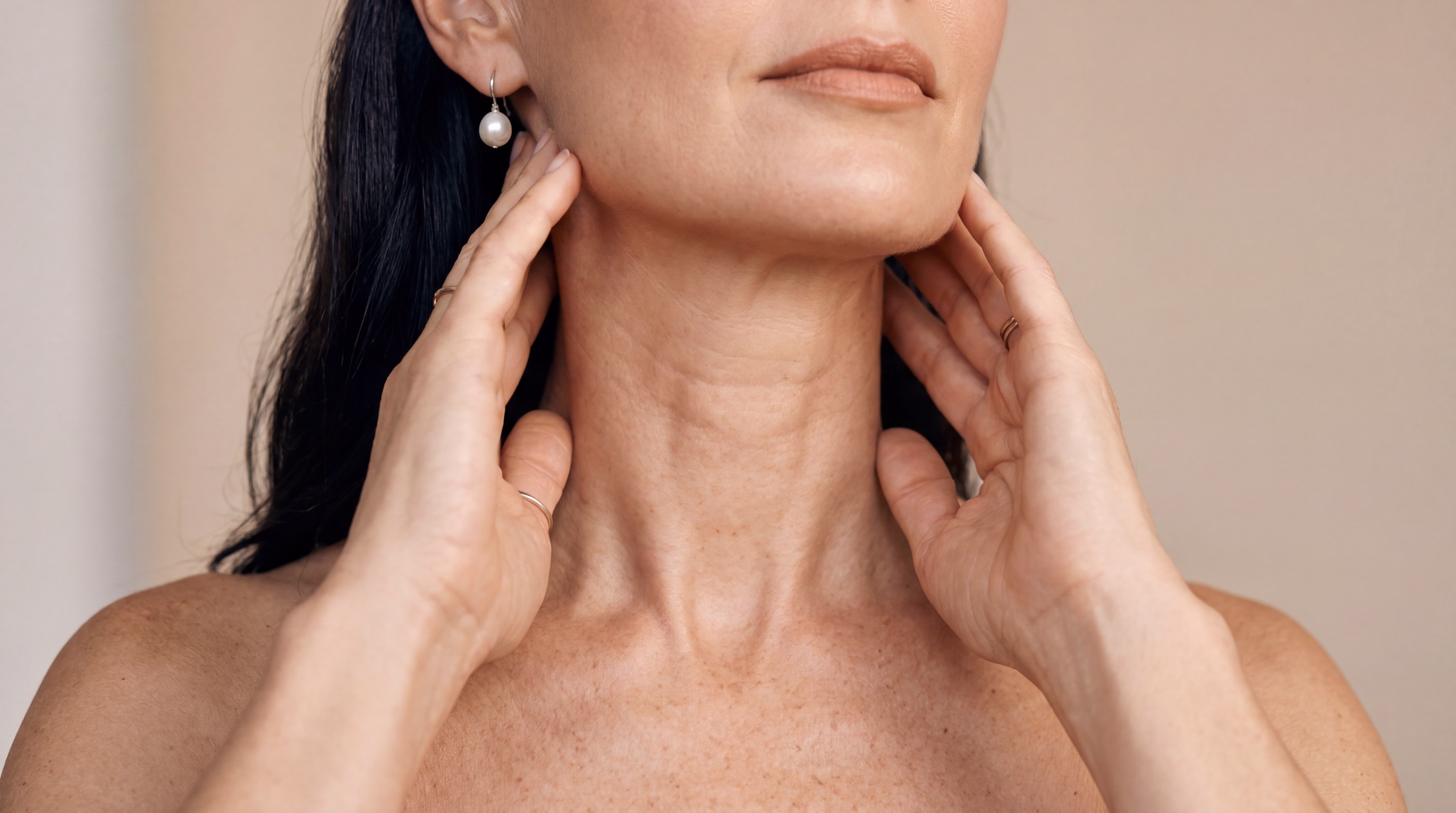 A woman gently touching her neck and jawline with both hands, wearing a pair of pearl earrings.