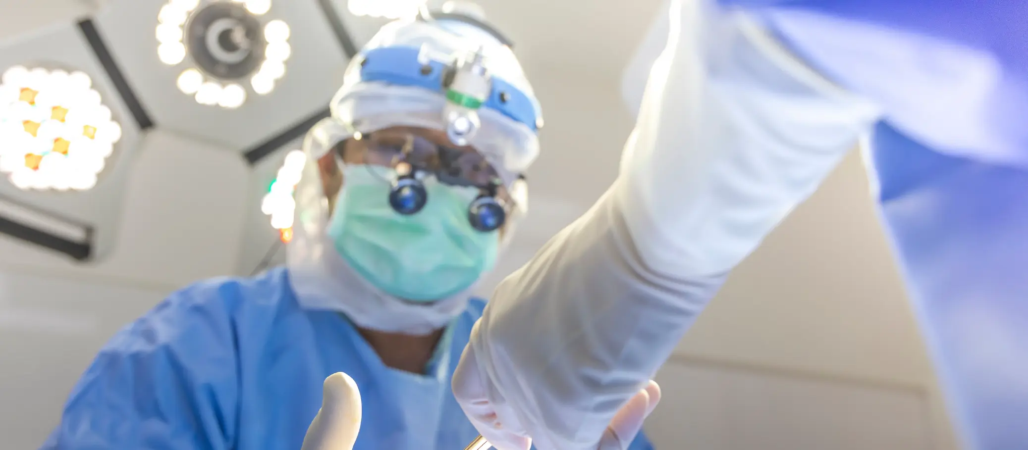 A medical professional wearing a surgical mask, protective eyewear, and surgical gloves is reaching towards a patient during a procedure, with overhead surgical lights illuminating the scene.