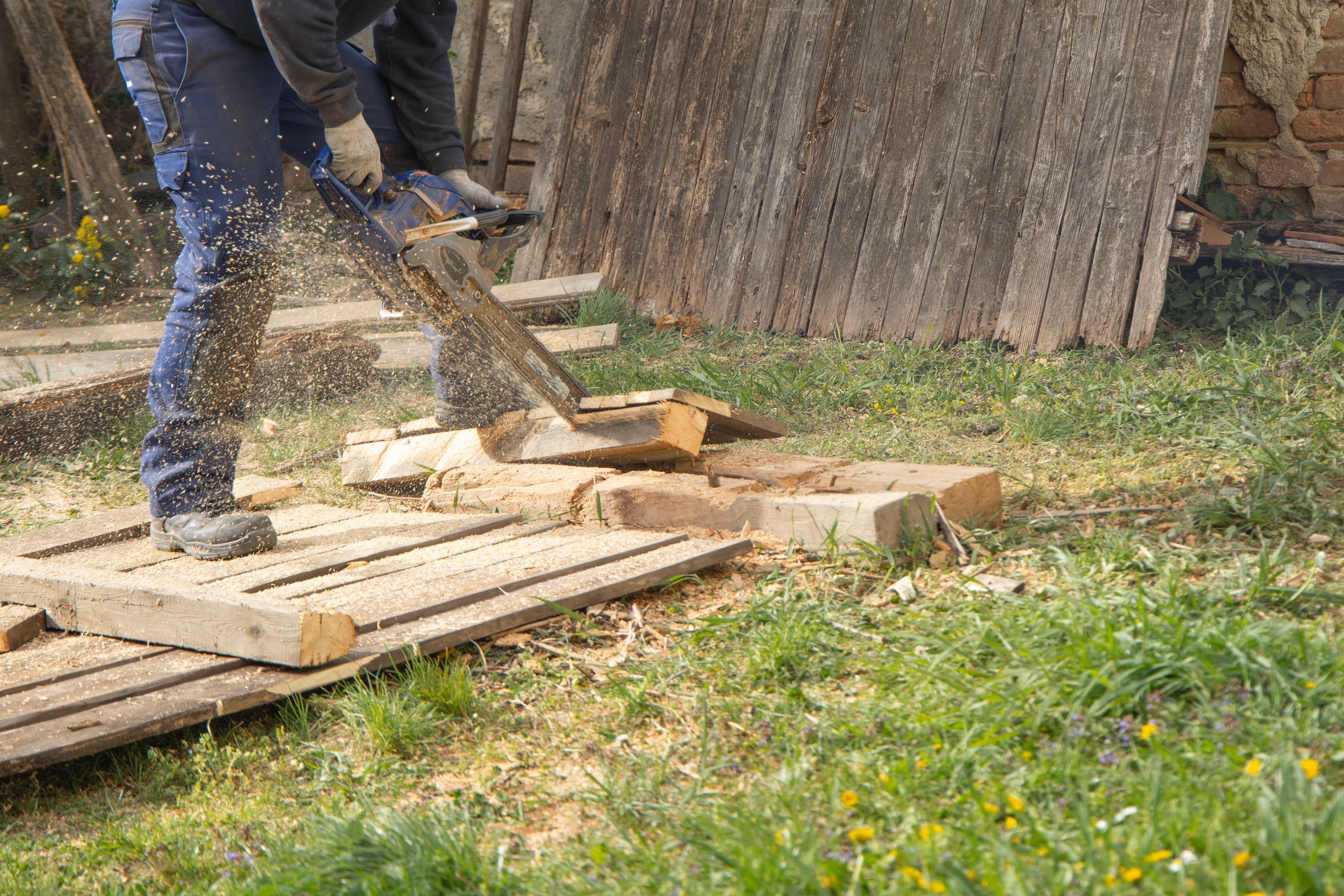 Worker using a chainsaw to cut and remove old wooden fencing outdoors, with sawdust flying around the work area.