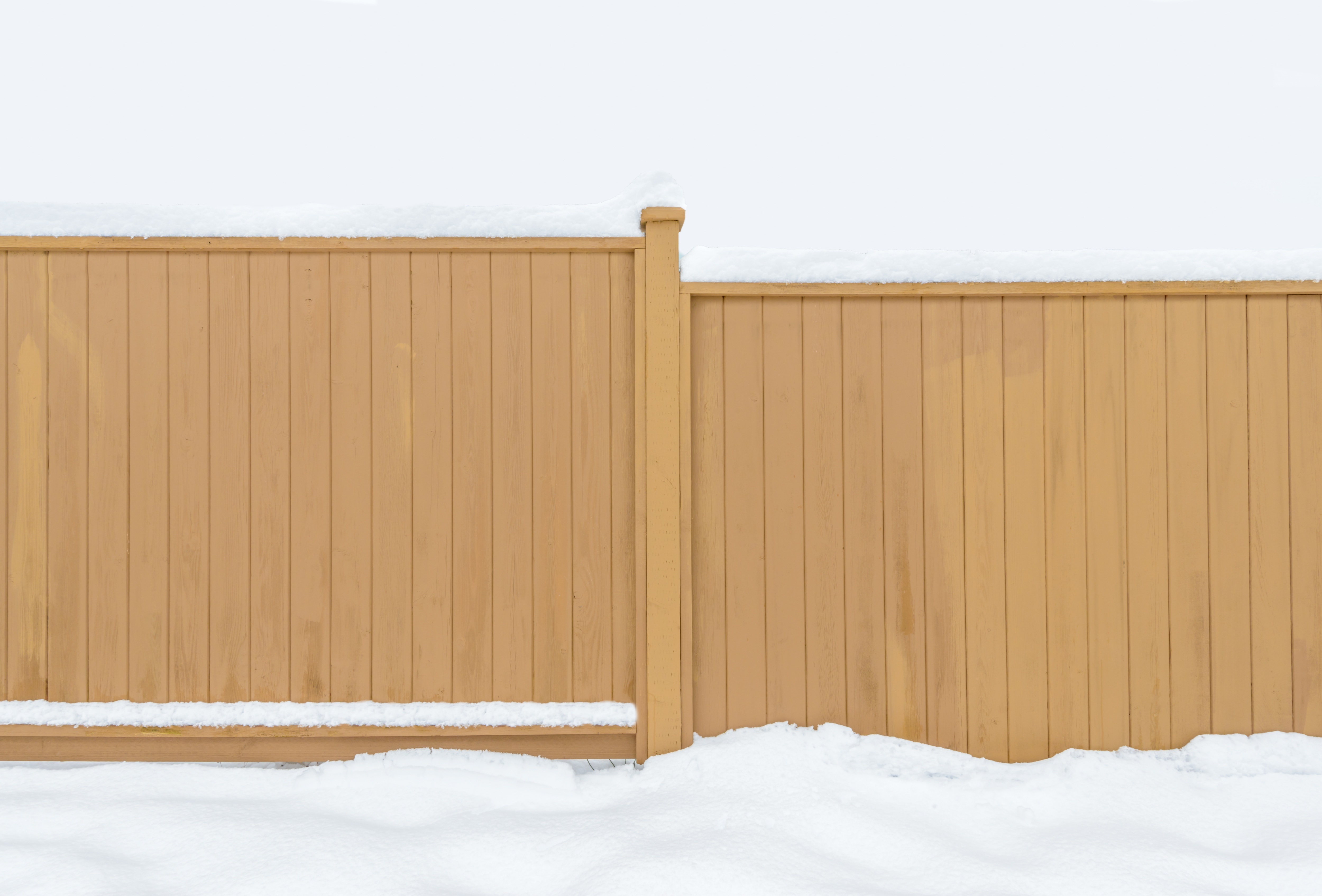 Light brown wooden privacy fence covered with snow, standing against a snowy white background in winter.