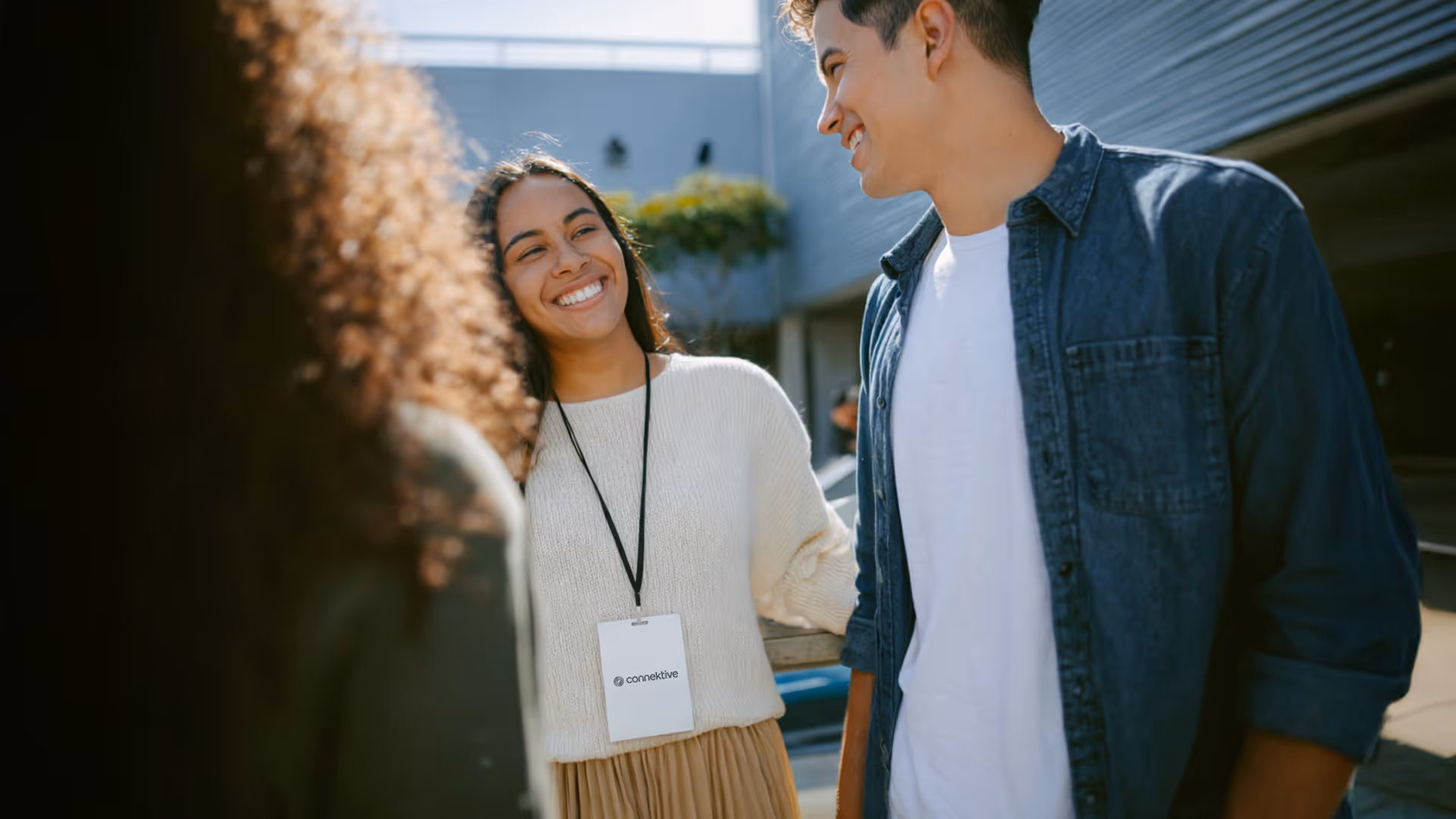 Smiling young woman wearing a white sweater and a Connektive badge talking to a young man in a denim shirt outdoors.