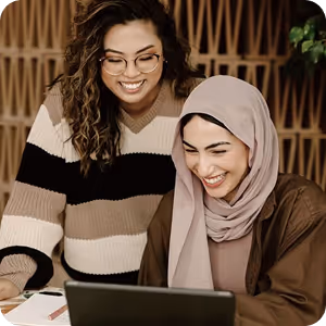 Two women smiling and looking at a laptop, one wearing glasses and a striped sweater, the other wearing a hijab and brown jacket.