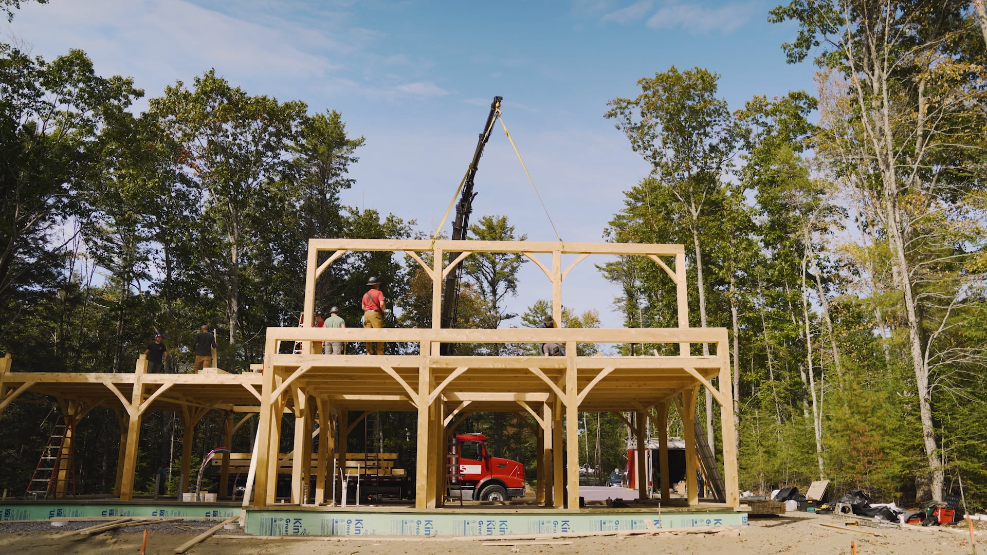 Wooden frame construction in progress with workers and crane in wooded area