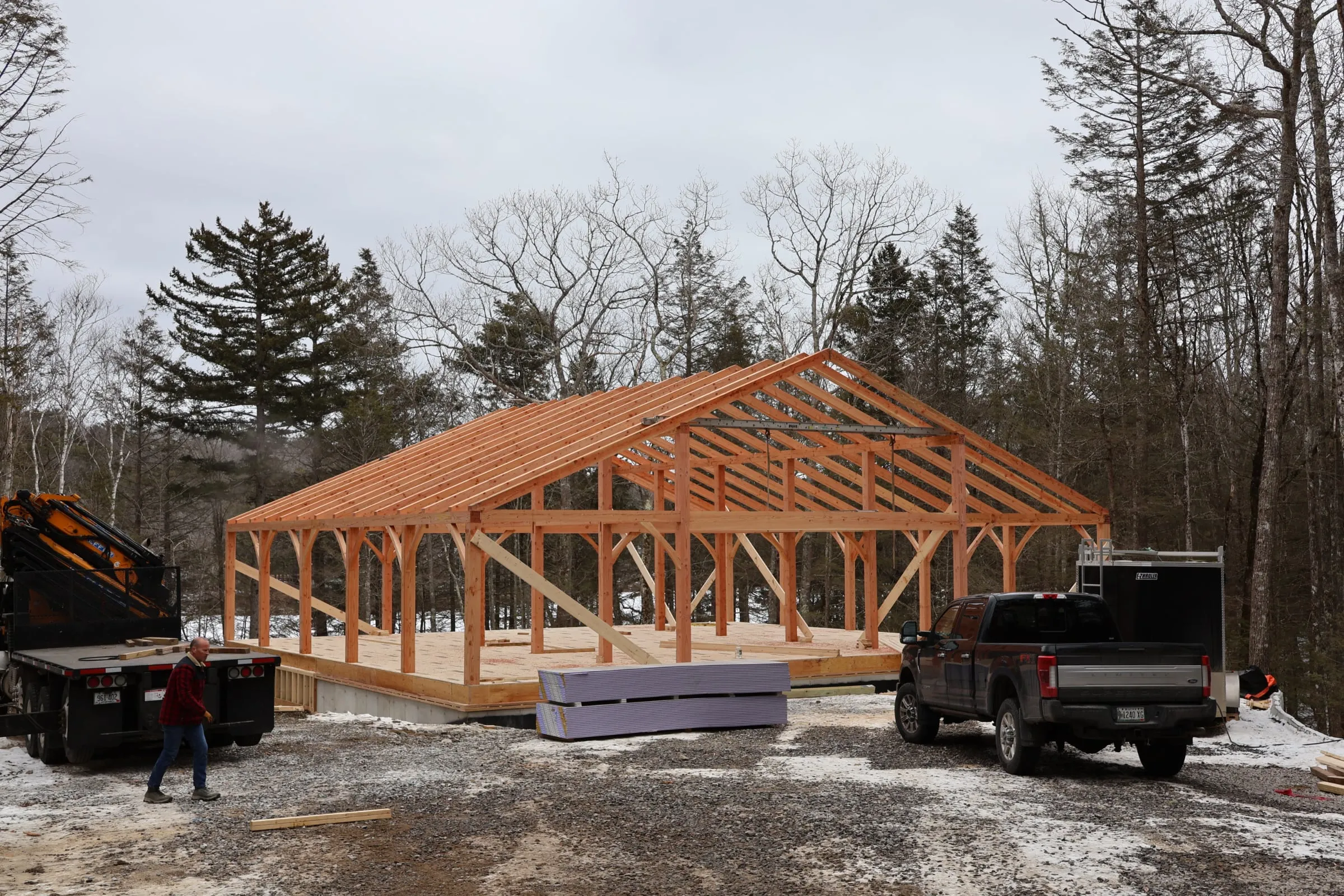 Wooden timber frame barn structure under construction in forest clearing with vehicles.