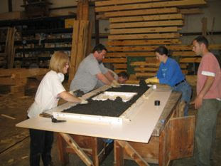 Four people examining architectural blueprint or floor plan on large table in woodworking workshop.