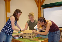 Four people collaborating on a wooden model house project at a table indoors.