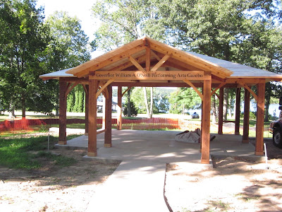 Large wooden pavilion with peaked roof in park surrounded by green trees.