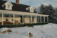 White farmhouse with black roof and wraparound porch in winter snow