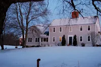 Pink colonial house in snowy landscape with bare trees at dusk.