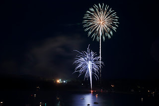 Gold and blue fireworks burst over dark water at night with distant lights
