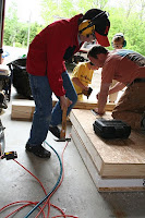 Man in red shirt using power drill on wooden workbench in garage.