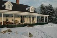 White farmhouse with black roof and wraparound porch in winter snow