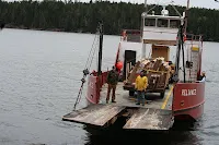 Red and white industrial barge with excavator on water near forested shoreline