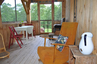 Modern cabin interior with wooden furniture, large windows overlooking green forest landscape outside.