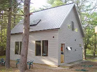Small gray cottage with metal roof surrounded by tall pine trees in forest setting