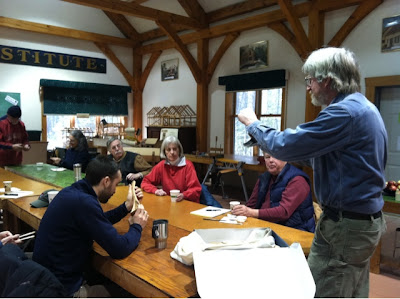 Elderly man presents to group of seated adults in wooden hall structure.