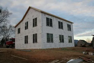 Two-story white house under construction with bare ground and trees surrounding it