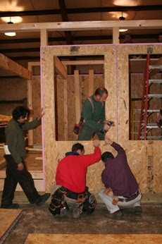 Construction workers building wooden frame structure indoors with tools and materials visible.