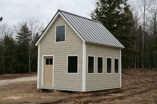 Small white cottage with metal roof and multiple windows surrounded by bare trees