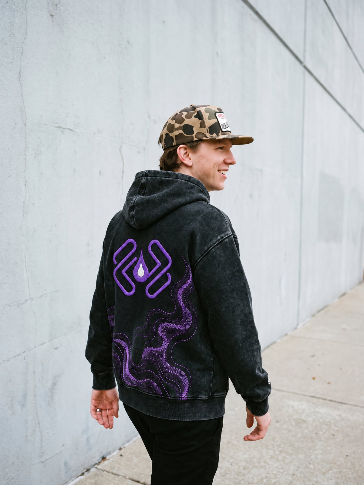 A man in a camo cap walking past a concrete wall, the purple back emblem of the Git Pullover toward the camera