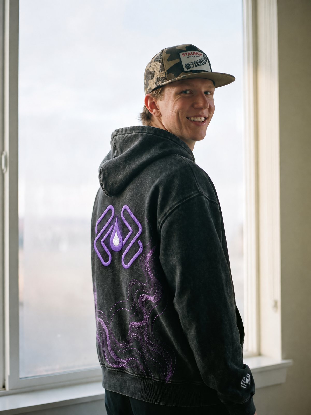 A man in a camo cap by a window looking over his shoulder, the back of the Git Pullover toward the camera